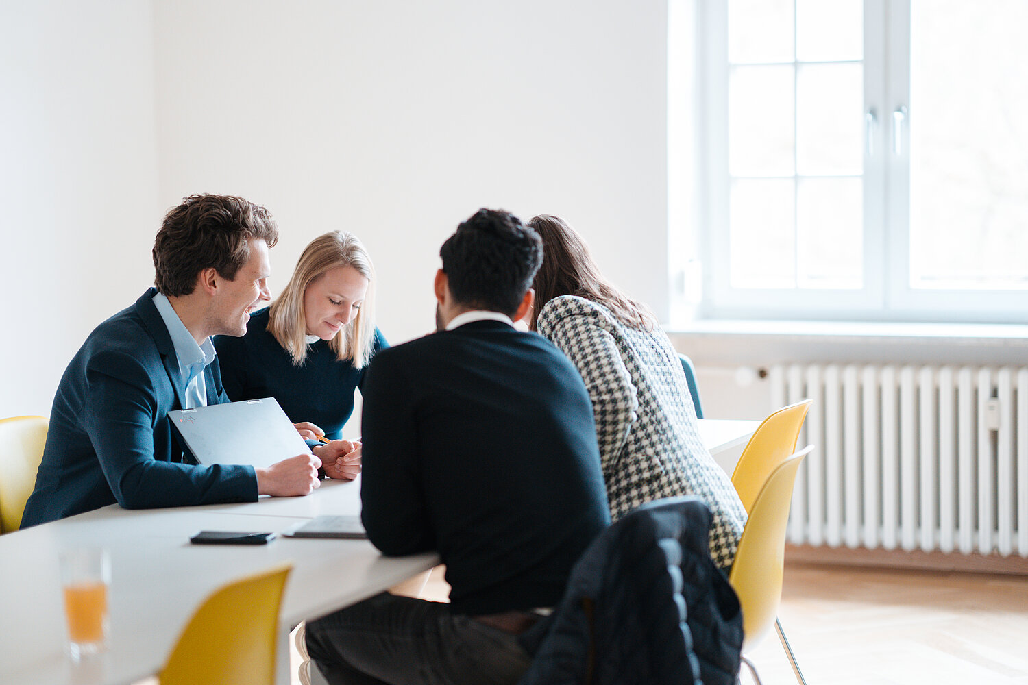 Vier Personen arbeiten gemeinsam am Tisch in einem hellen Büro. Zwei sehen sich Notizen an, während die anderen zuhören. Stühle sind gelb, Fenster im Hintergrund.