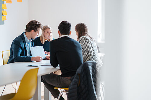 Personen in einem Meeting am Tisch, fokussiert und diskussionierend, mit Notizzetteln an der Wand im Hintergrund und warmem Tageslicht im Raum.