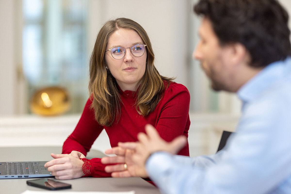 Zwei Personen in einer Besprechung, Frau in rotem Pullover mit Brille konzentriert zuhörend, unscharfer Mann im Vordergrund gestikulierend, Bild bei undconsorten.de.
